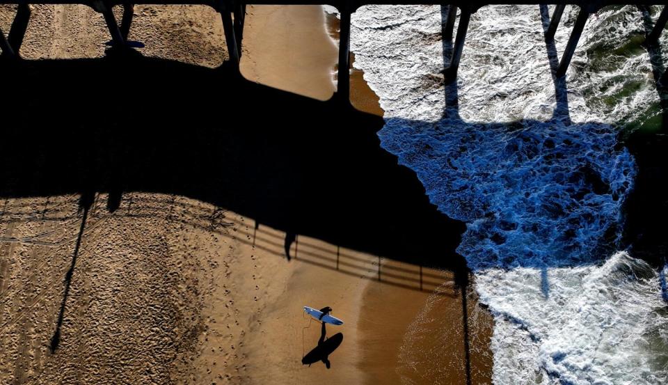 The morning sun creates shadows from the pier across the sand and water and a surfer heading out.