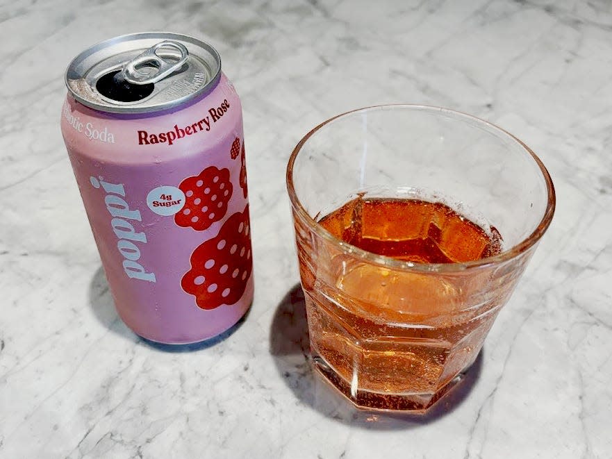 An open can of raspberry rose Poppi next to a small, clear glass with pink liquid inside. Both are sitting on a countertop.