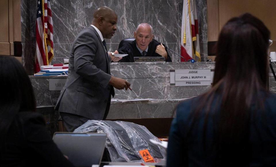 Evidence, including the clothing of victim Anthony Williams, sits at the front of the court room while David Howard, defense attorney for Jamell Demons, better known as YNW Melly, hands a paper to Judge John Murphy during the trial on Tuesday, June 20, 2023, at the Broward County Courthouse in Fort Lauderdale, Fla.
