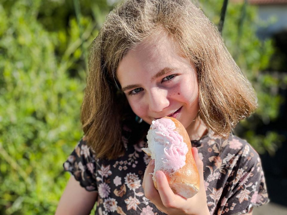 A girl smiles and holds a raspberry bun, which looks similar to a long John. The girl wears a black top with a pink floral print.