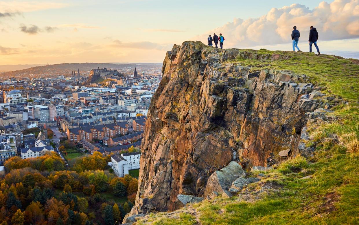 Group of people walking along cliff edge looking at city views. Edinburgh Castle in the distance