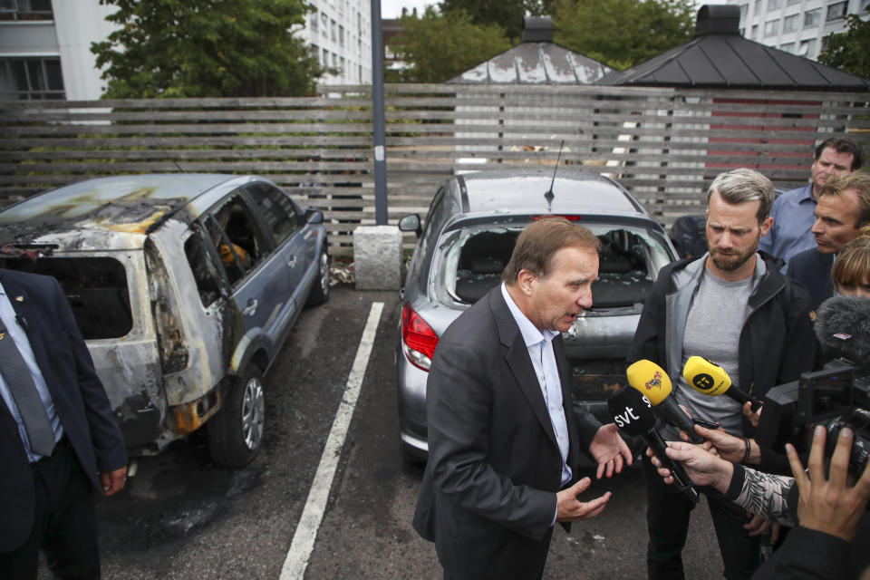 Swedish Prime Minister Stefan Lofven foreground, talks to journalists, after cars were set on fire, at Frolunda Square, in Gothenburg, Sweden, Tuesday, Aug. 14, 2018. Masked youth torched dozens of cars overnight in Sweden and threw rocks at police, prompting an angry response from the prime minister, who spoke of an "extremely organized" night of vandalism. (Henrik Brunnsgard/TT News Agency via AP)