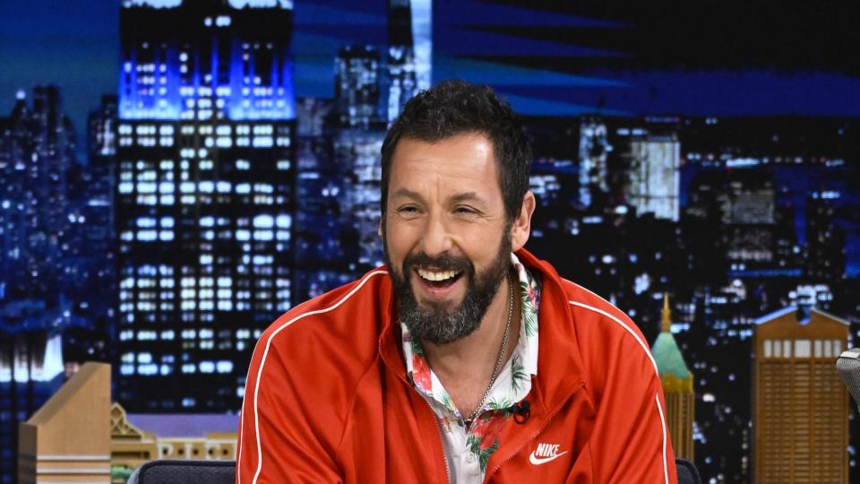 adam sandler smiling and looking toward the audience while sitting for a talk show appearance