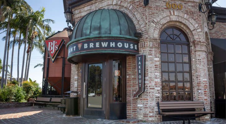 An image of the entrance to a brick BJ's Restaurant and Brewhouse building with a green auning, wood door, bench, and brick walkway.