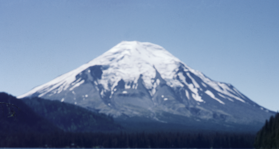 A blurry photograph of a snow-capped mountain with forests at its base. No visible persons or text