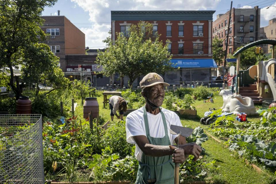 A man with hoe stands in front of the community farm with play equipment to one side and buildings in the background.