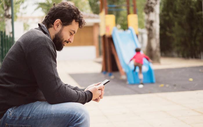 A man with a beard sits on a park bench looking at his phone. A child in a red shirt is playing on a slide in the background