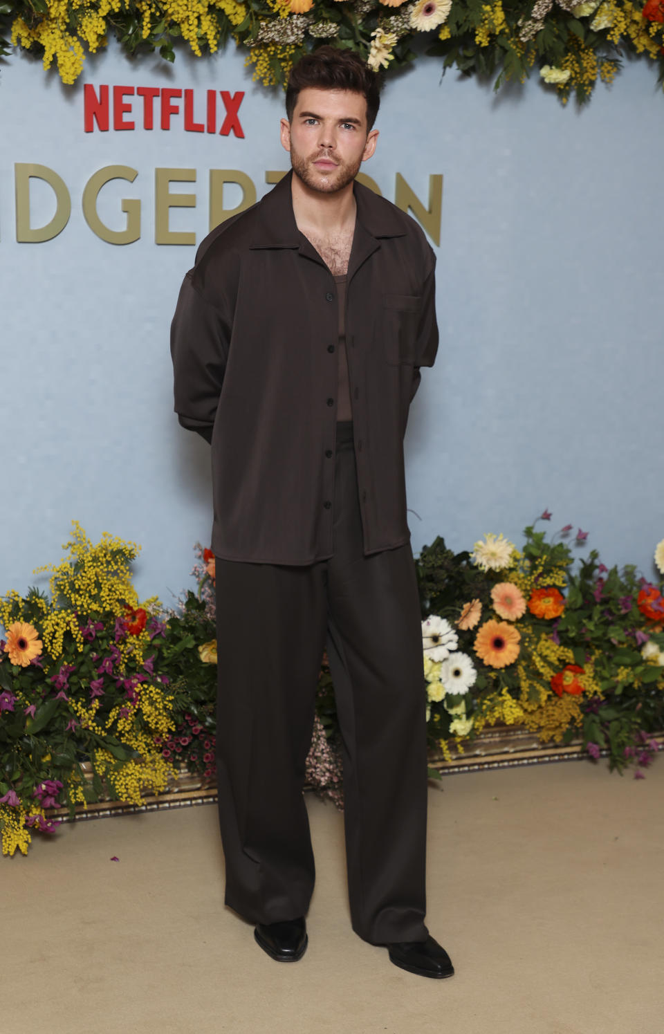 Man stands in front of floral backdrop wearing an oversized brown shirt and matching trousers