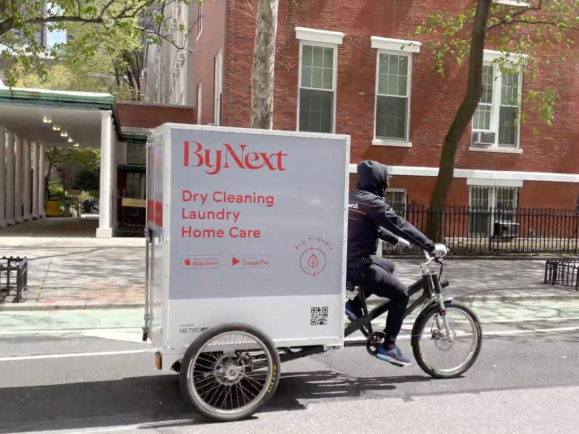 A delivery worker rides a cargo bike with a trailer that says "ByNext" down a street with brick buildings in New York City.