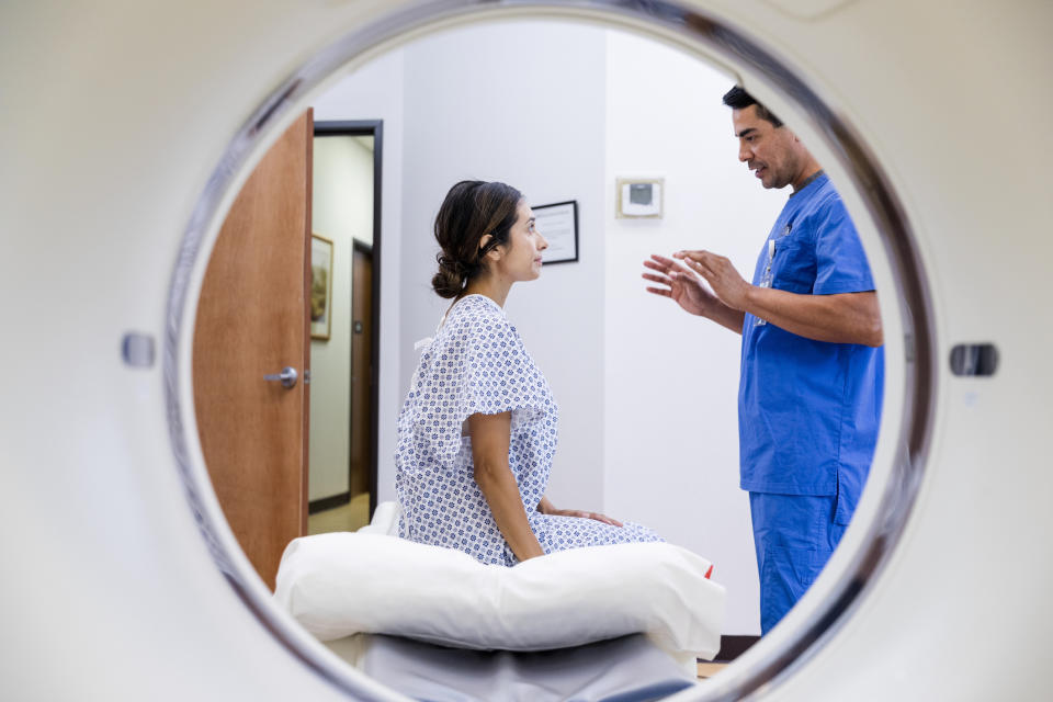 A woman in a hospital gown sits on a medical bed inside an MRI machine, speaking with a male doctor in blue scrubs