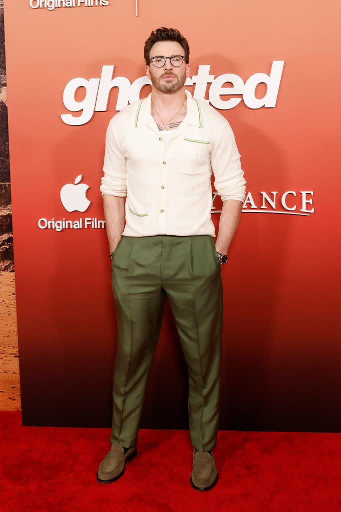 Man in glasses poses in a cream cardigan, green pants, and matching shoes at a film event