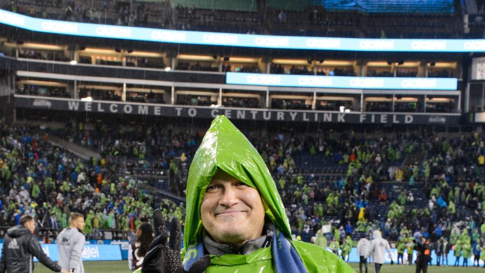 drew carey waving for a photo while wearing a scarf and rain poncho