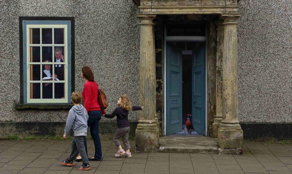 A family walks past an empty building, which has been covered with artwork to make it look more appealing, in the village of Bushmills on the Causeway Coast August 20, 2013. One of the homes of Irish whiskey has taken a scheme developed in Northern Ireland of erecting fake shop fronts where derelict buildings lie and has truly run with it in a bid to woo tourists. Bushmills, best known as the village where the whiskey of the same name was distilled for the first time 400 years ago, is now also becoming recognisable for the artwork and graphics that brighten up shop fronts left empty during the economic downturn. Picture taken August 20, 2013. REUTERS/Cathal McNaughton (NORTHERN IRELAND - Tags: BUSINESS SOCIETY TRAVEL) ATTENTION EDITORS: PICTURE 01 OF 20 FOR PACKAGE 'NORTHERN IRELAND'S TROMPE L'OEIL' SEARCH 'BUSHMILLS ART' FOR ALL IMAGES