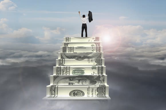 A man celebrating after climbing to the top of a staircase made out of money overlooking the clouds with a brightly shining sun.