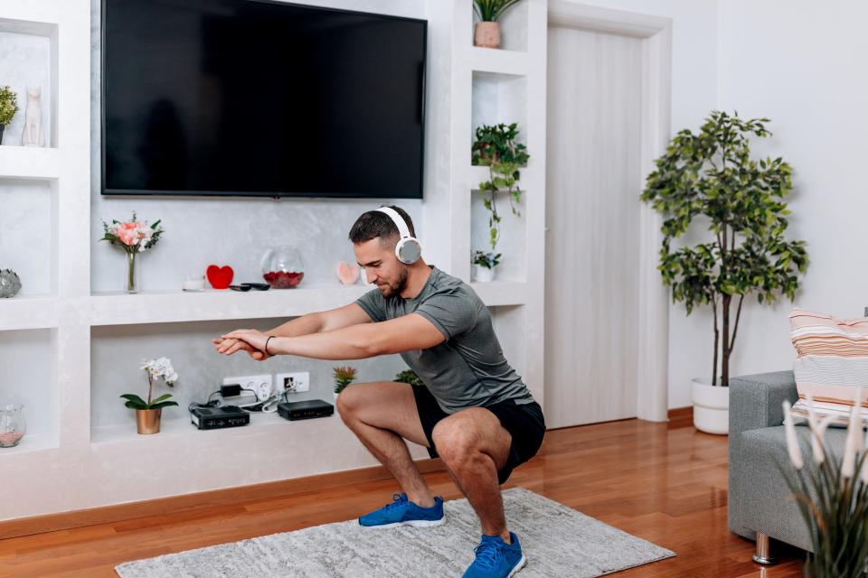 A man holding a deep squat position in while wearing a t-shirt, shorts, and headphones in his living room.