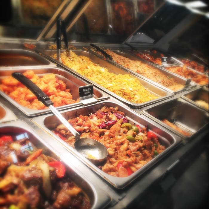 Buffet with various dishes including stir-fry, pasta, rice, and fried food in metal trays with serving utensils