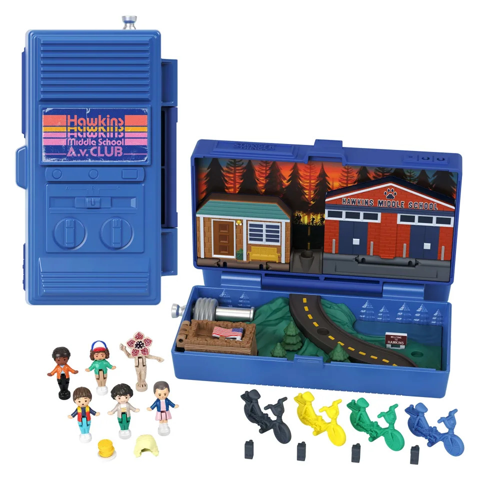 Includes The Stranger Things Polly Pocket set and figurines displayed on a white background. It has a blue wireless shaped compact shown closed on the left and open on the right. On the open side, Hawkins has representation, including high school. There are figurines for Eleven, Mike, Will, Lucas, Dustin and the Demogorgon, plus mini radios and bikes.