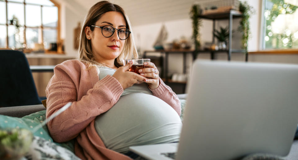 A pregnant woman sitting down on a couch with a laptop on her thighs. Source: Getty 