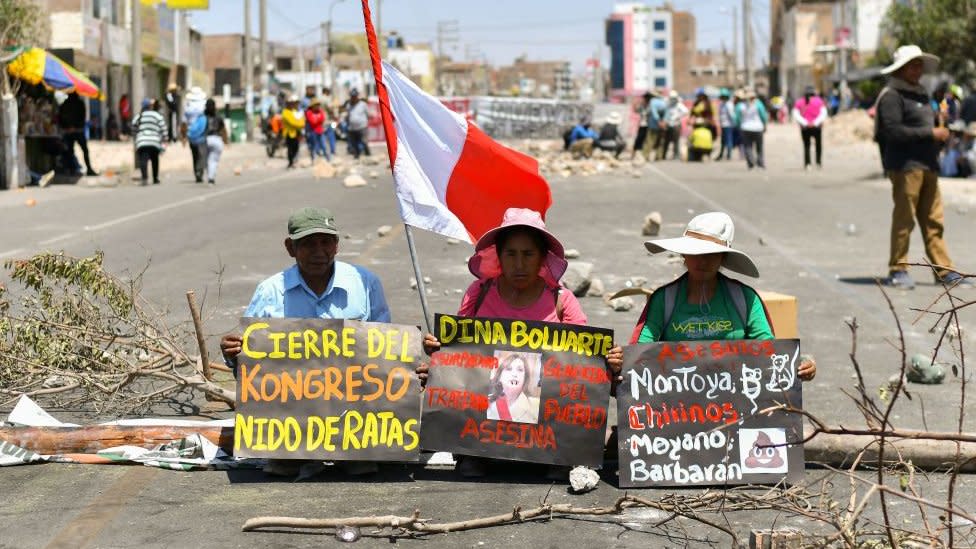 Manifestantes sentados con pancartas cortan el paso en una carretera peruana.