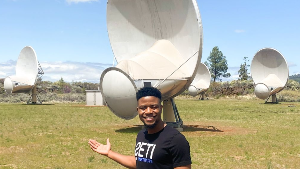  ramiro saide stands in front of a set of radio telescopes in a field with blue sky in behind 