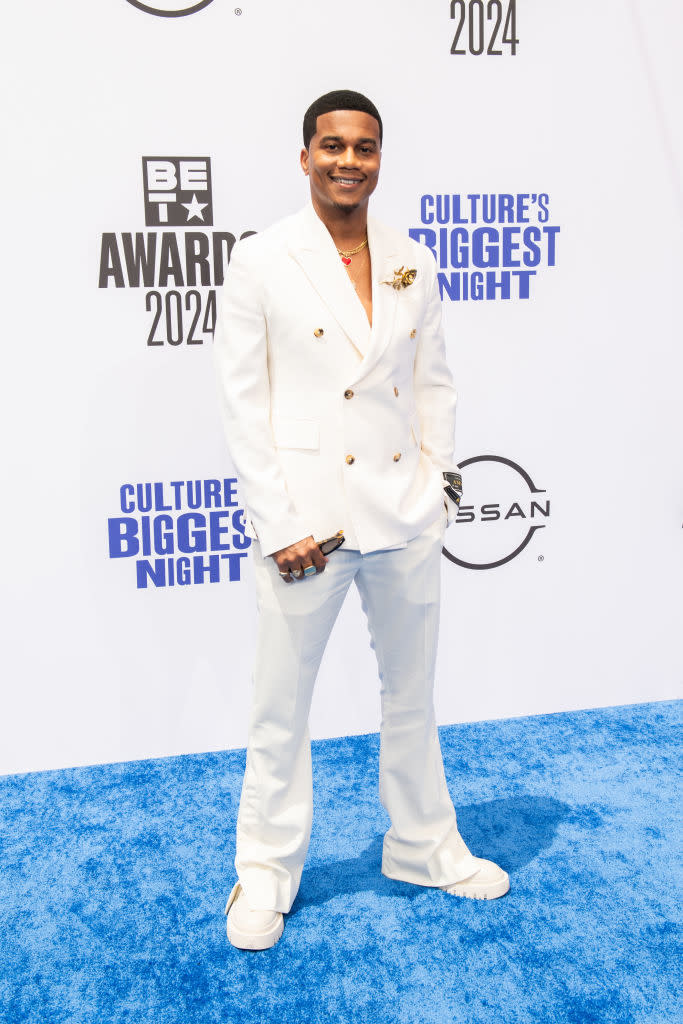 Cory Hardrict smiles on the red carpet, wearing a white double-breasted blazer, matching trousers, and white shoes, at the BET Awards 2024 event