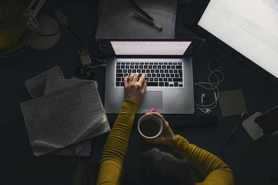 Overhead view of a person using a laptop with a notepad and a cup in hand, on a desk with various items