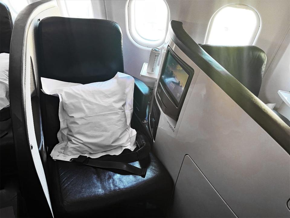 A lavender pillow on an airplane seat with a white panel with storage space and a screen next to it. A tray with a can of water on it is in the background