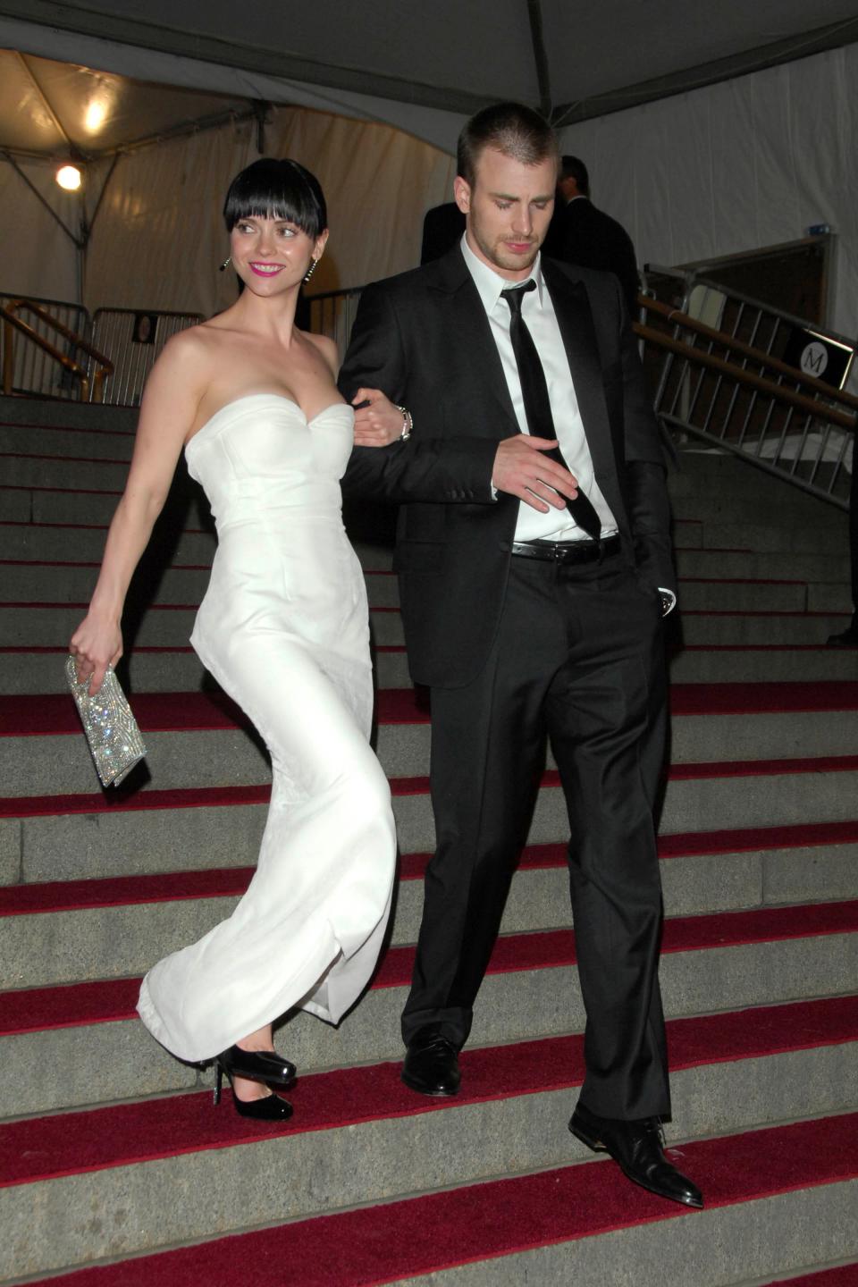 Christina Ricci wears a white dress and walks down a flight of stairs with Chris Evans, who wears a black suit.