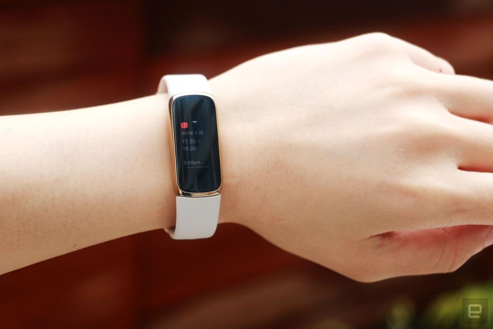 <p>Full frontal view of the Fitbit Luxe with a light pink silicone band on a wrist against a dark brown background. The screen shows a calendar notification for an event from 5:30pm to 7pm.</p>
