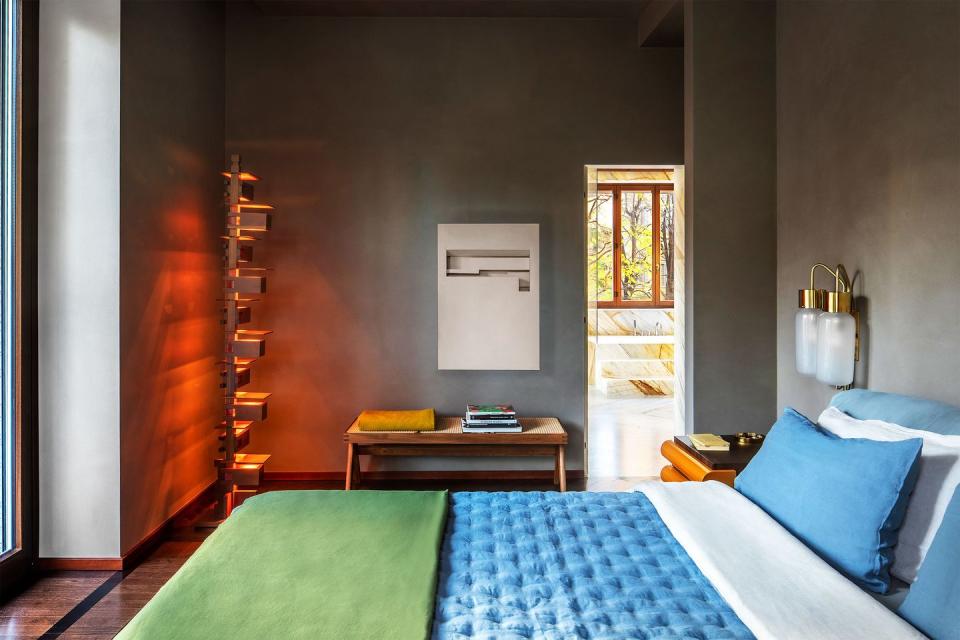 a bedroom with gray walls, bed with blue upholstered headboard with sconces above, blue pillows and bedcover, green blanket at foot, multilevel floor lamp with red tinted light, side table with magazines