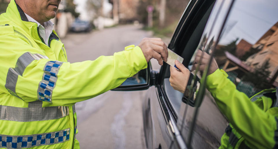 A police officer in a high-vis jacket hands a licence back to a driver of a black car.