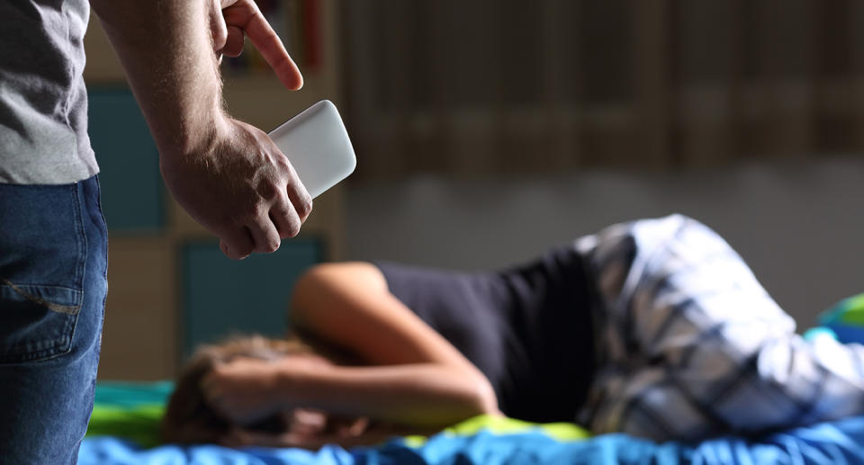 Father pointing mobile phone after discover bad content to a sad teen who is lying on the bed with a dark light in the background. Source: Getty Stock