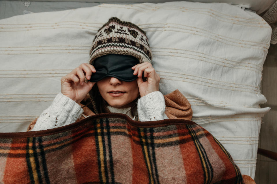A young woman in a warm hat and sweater lifts a sleeping mask while in bed. (Getty Images)
