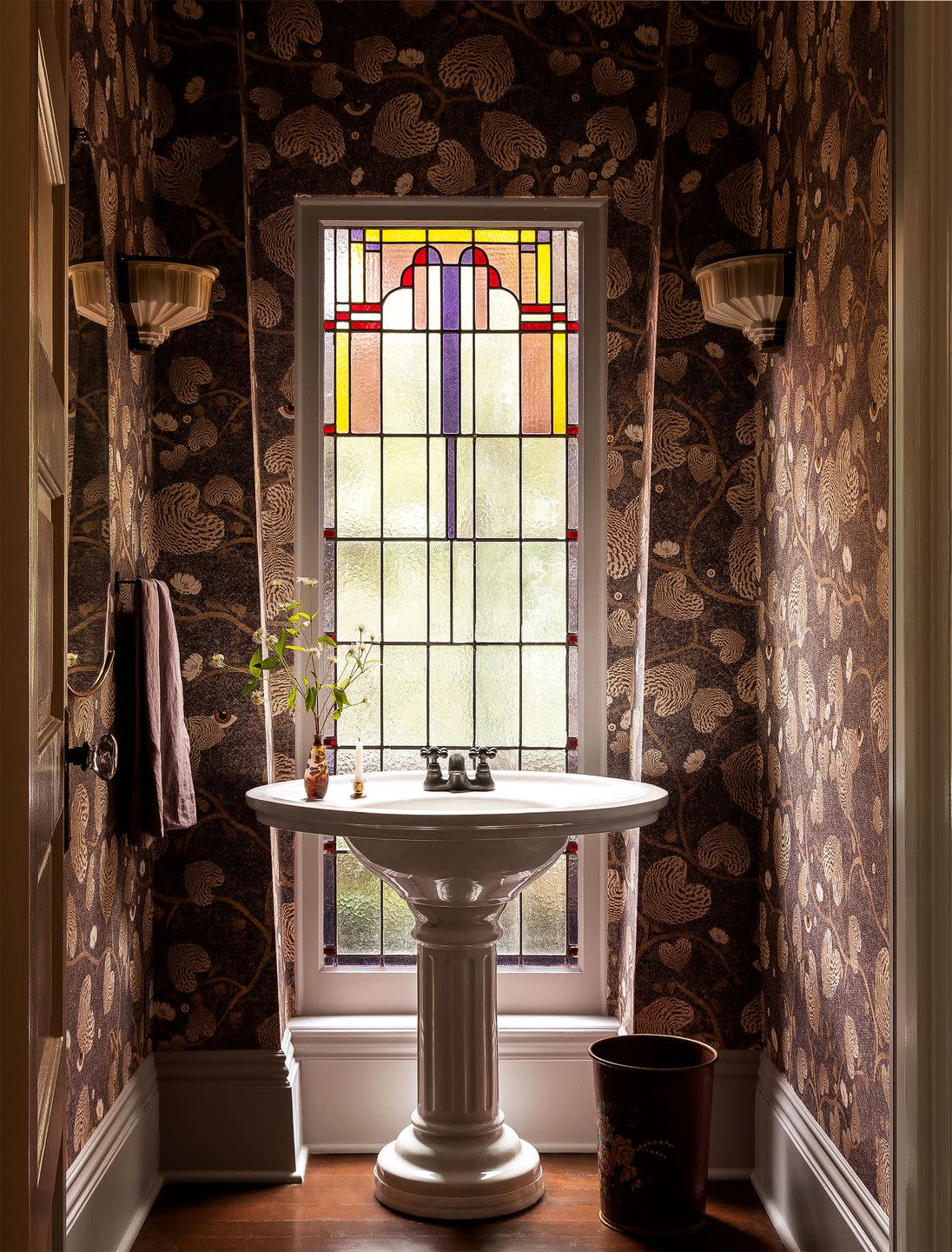 powder room with a brown background leaf pattern wallpaper, vintage sconces, a pedestal sink in front of a tall window with stained glass
