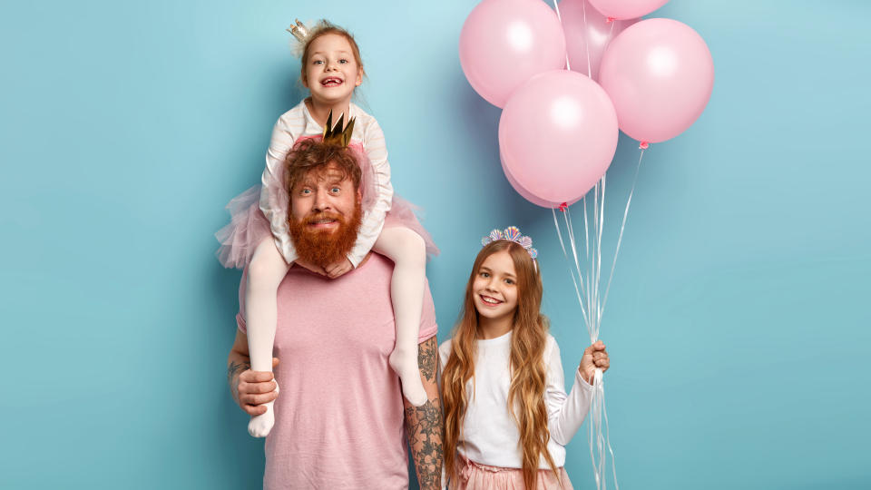 Studio shot of caring father gives piggyback to small daughter, little girl carries air balloons, celebrate birthday, spend free time at home and have fun, isolated over blue background. Ginger family