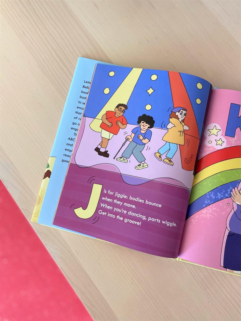 Photo from above of a page in the book, "B is For Bellies." The page shows three people dancing under a yellow and red spotlight and reads, "J is for jiggle; bodies bounce when they move. When you're dancing, parts wiggle. Get into the groove!" 