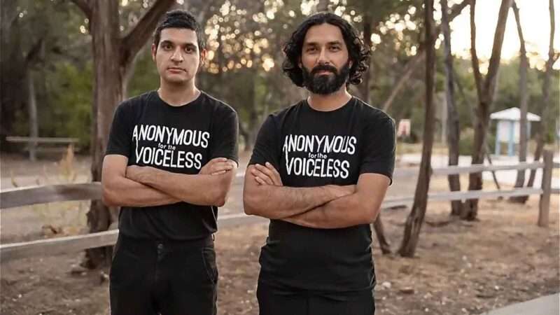 two animal rights protesters, Daraius Dubash and Faraz Harsini, stand arms-crossed in a park wearing "Anonymous for the Voiceless" T-shirts.