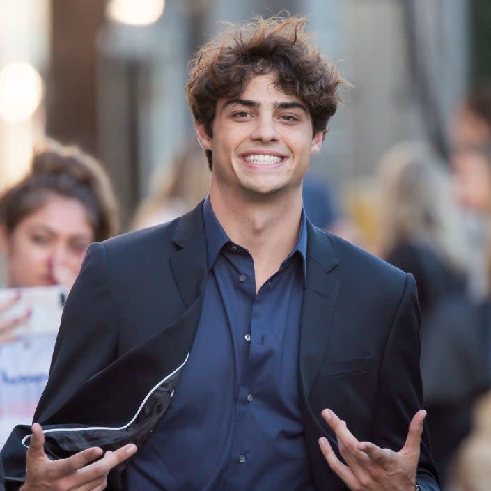 Person in a black suit without a tie, smiling and gesturing with hands up