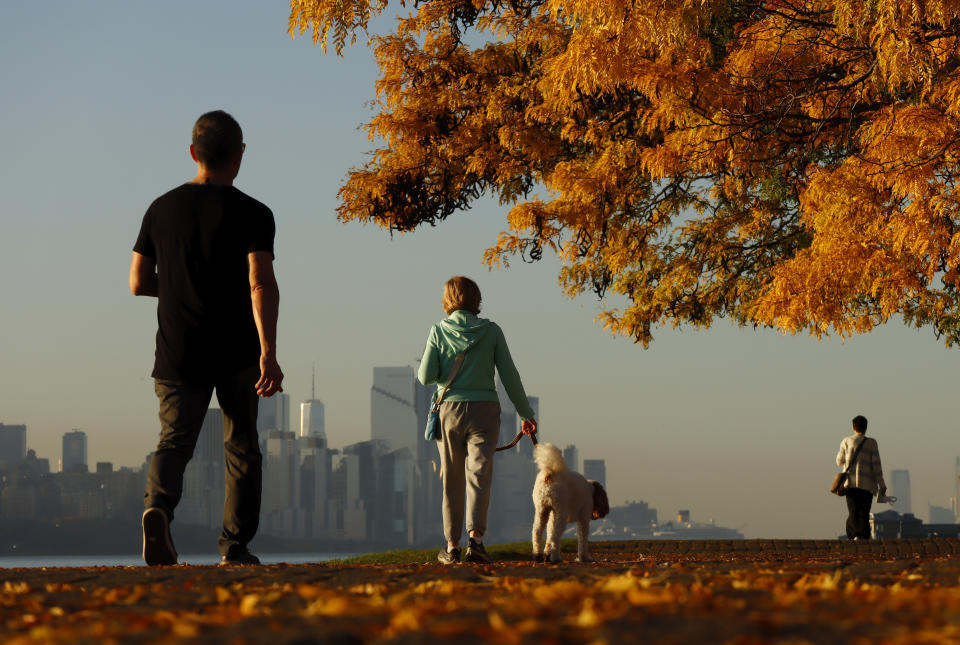 People walk along a walkway covered in fall foliage in front of Hudson Yards and One World Trade Center in New York City as the sun rises in October 2023, in Edgewater, New Jersey.