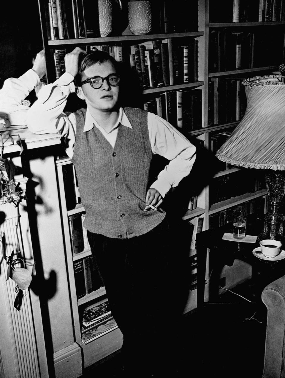 truman capote looks at the camera while standing in front of a book shelf, he leans his head against one arm propped up on a mantle as he holds a cigarette in his other hand, he wears a sweater vest, collared shirt, pants, and glasses