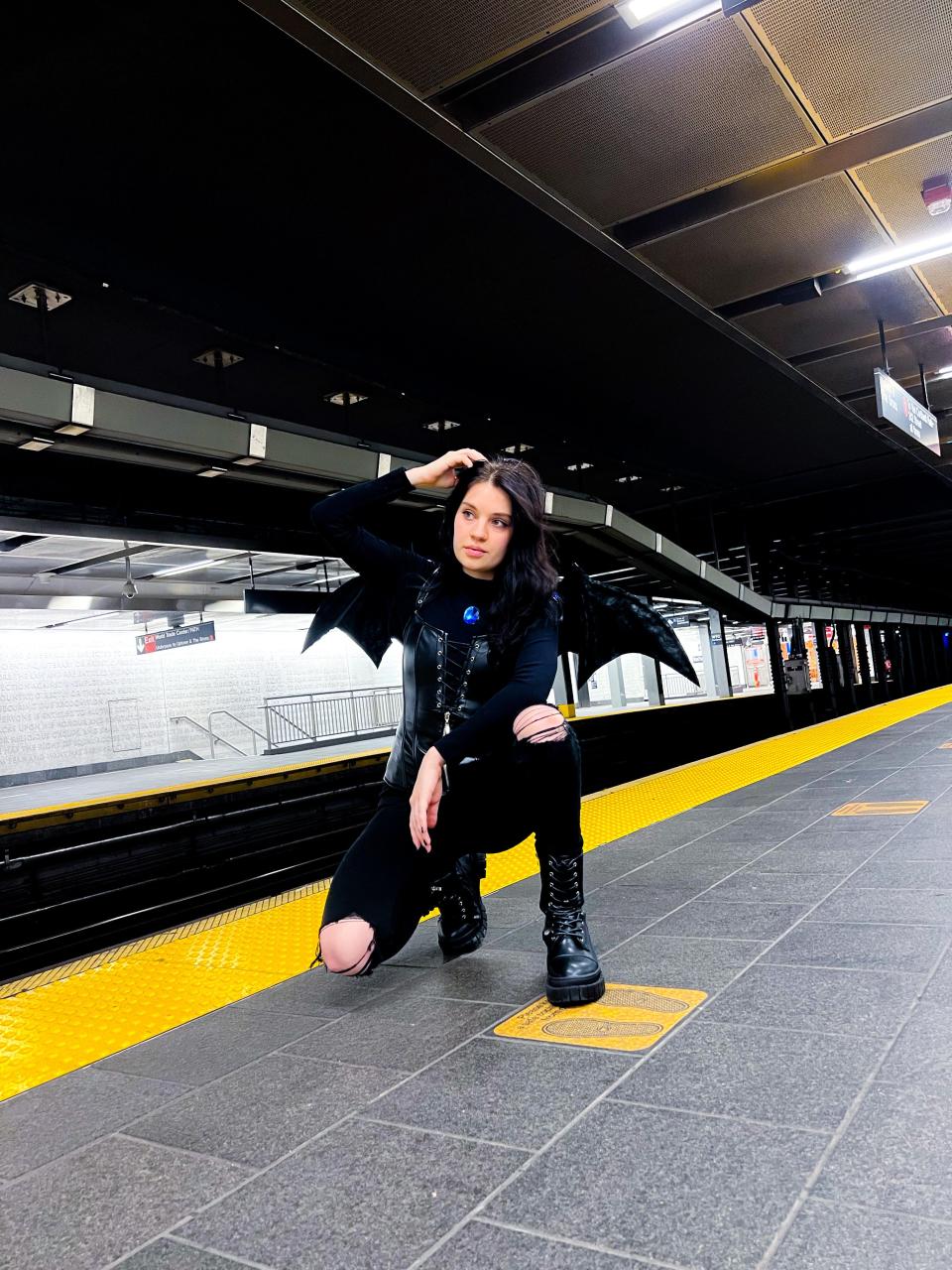 A woman in a costume with bat wings stands in a train station.