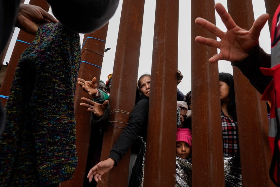 FILE – Migrants reach through a border wall for clothing handed out by volunteers as they wait between two border walls to apply for asylum on May 12, 2023, in San Diego. President Joe Biden has ordered a halt to asylum processing at the U.S. border with Mexico when arrests for illegal entry top 2,500 a day, which was triggered immediately. (AP Photo/Gregory Bull, File)