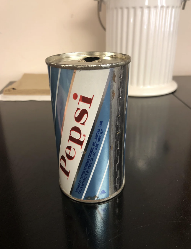 A vintage Pepsi can with a pop-top opening sits on a dark table. A white, ribbed container is blurred in the background