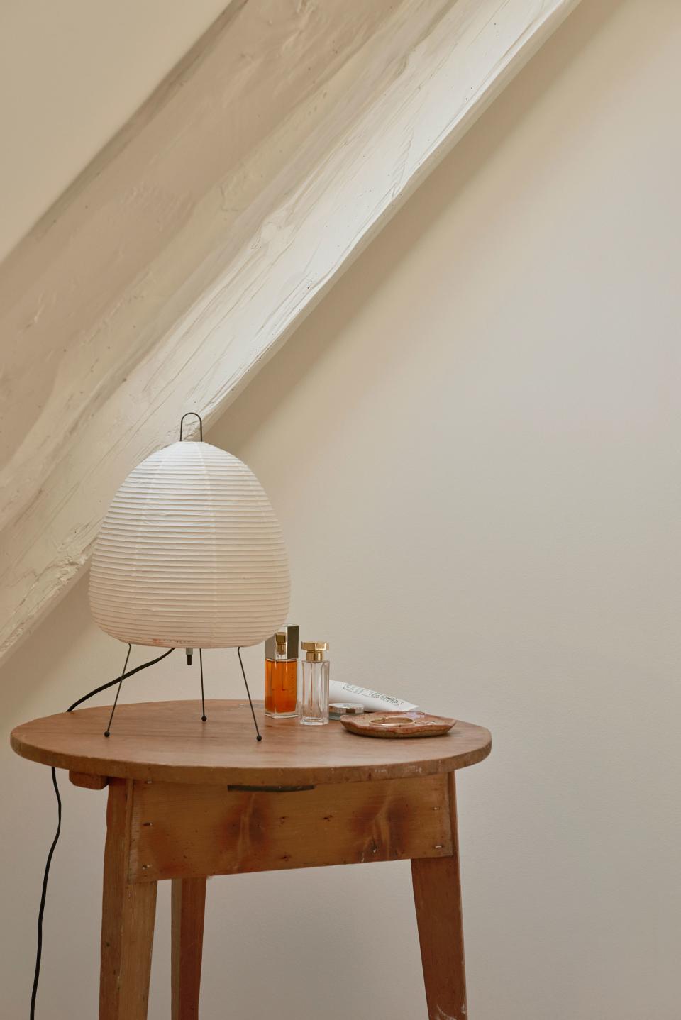 An Akari light fixture provides light atop an antique wood round table in the bedroom, all in front of one of the slanted wood beams that she painted white (they run all along one wall of every room in the apartment, adding character but also making the layout more complex).