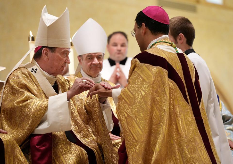 The Rev. Earl K. Fernandes, right, kneels before Archbishop Dennis M. Schnurr, left, and Bishop Robert J. Brennan, center, during his ordination and installation Tuesday as the 13th bishop of the Diocese of Columbus.