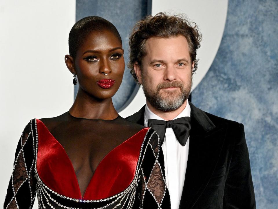 Jodie Turner-Smith, in a black and red sheer gown with rhinestone accents and matching red lipstick, poses on the red carpet with husband Joshua Jackson, who is wearing a black velvet tux and has his arm around Turner-Smith's waist.