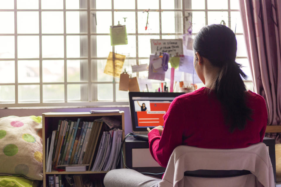 Asian Teenage girl wearing earphones sitting in front of lapttop, attending online video classroom with teacher in her bedroom