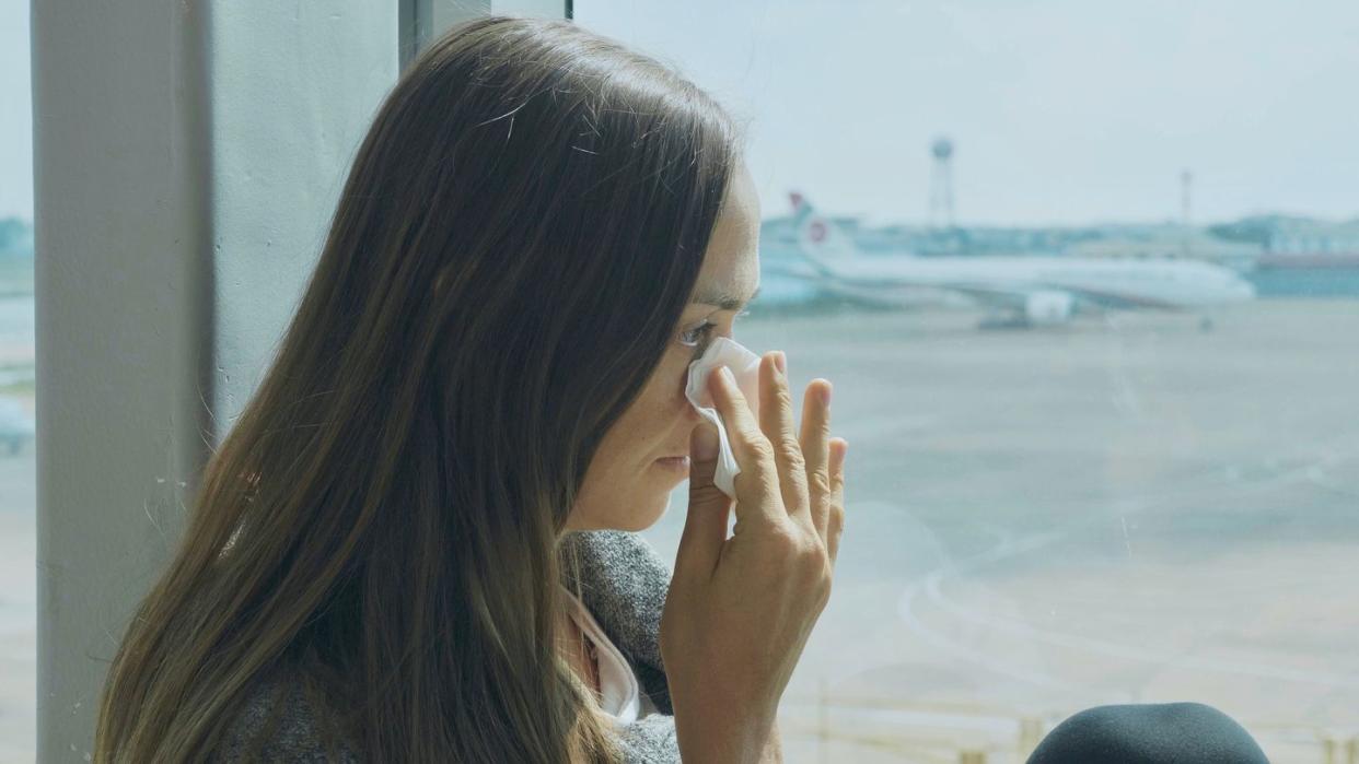 young sad woman is sitting on the window sill at airport, crying and wipes tears away with airplane on the background