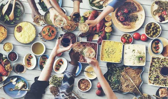 A table set with various foods. People off screen have their hands over the table toasting.