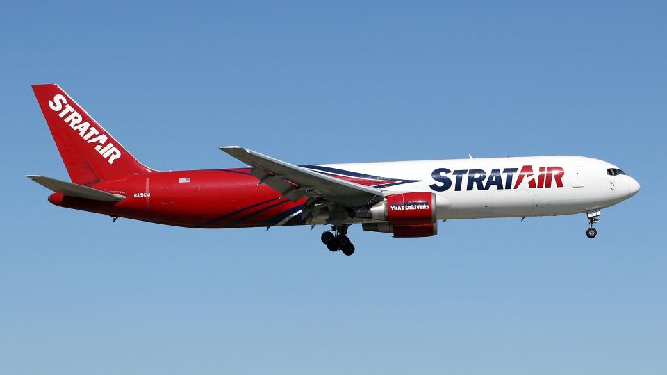 Half-red, half-white cargo jet with logo Stratair approaches airport with wheels down, as seen from below.
